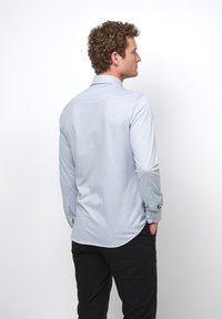 Camisa de manga larga azul claro con cuello botón, textura suave, dobladillo curvado y detalles de botones en los puños, combinada con pantalones negros.