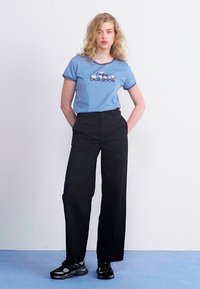Mėlyna medvilninė marškinėliai su "New Balance" tekstu, derinami su aukštu juosmeniu juodomis plačiomis kelnėmis ir juodais sportbačiais. Paprastas, kasdieniškas įvaizdis.