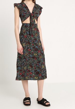 Kobieta w czarnej kwiatowej sukience midi z wiązaniem z przodu i wycięciem, zestawiona z czarnymi plecionymi klapkami na jednolitym tle.