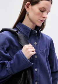 Τζιν μπουφάν σε ναυτικό μπλε με κούμπωμα μπροστά, μεγάλο γιακά και υφασμένα μανίκια· συνδυασμένο με τσάντα με μαύρο δερμάτινο λουράκι. Ασημί κουμπιά ως διακοσμητικά στοιχεία.