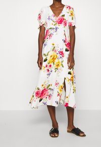 Blommig midiklänning med V-ringning, vit bas, korta puffärmar, stora rosa och gula blommor samt sidoklyv. Kombinerad med svarta sandaler.