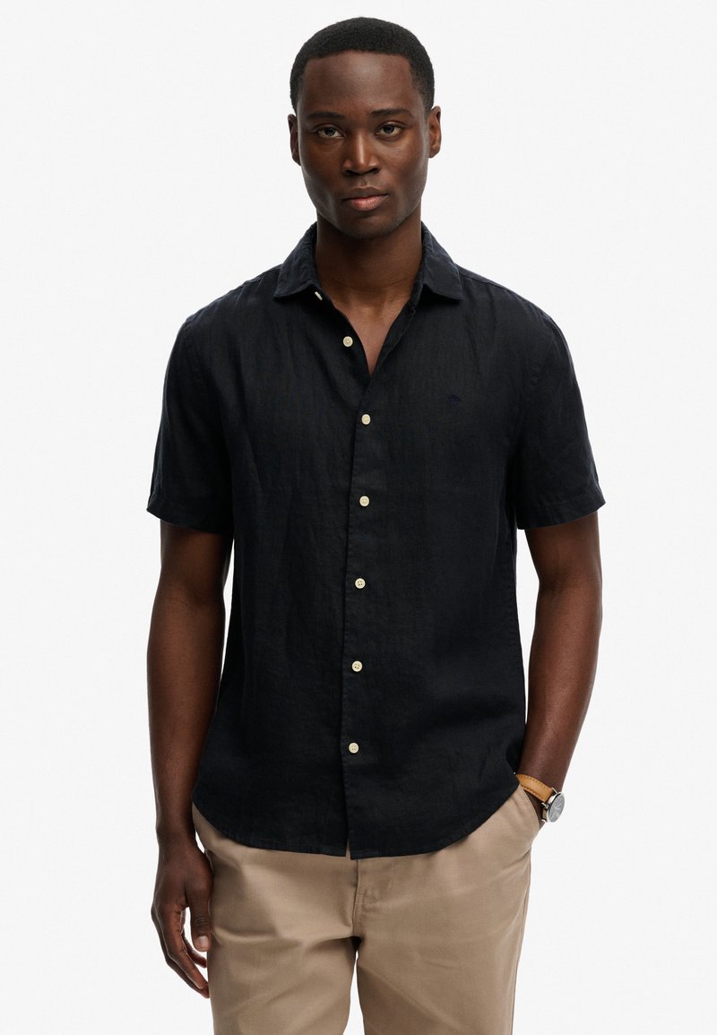 Homme portant une chemise noire à manches courtes boutonnée et un pantalon beige, debout avec une main dans la poche devant un fond uni.