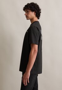 Zwarte katoenen t-shirt met korte mouwen en een ronde hals. Bevat tekst op de voor- en achterkant. Draagt bij donkere spijkerbroeken, gezien vanaf de zijkant.