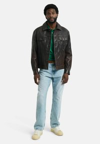 Braune Lederjacke mit zwei Fronttaschen, klassischem Kragen und Knopfverschluss, kombiniert mit einem grünen Poloshirt und hellblauen Jeans.