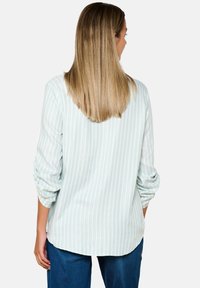 Blusa a righe verde chiaro con maniche lunghe, dotata di un orlo arrotondato e una trama liscia. La vista posteriore mostra le maniche arrotolate e una vestibilità rilassata.