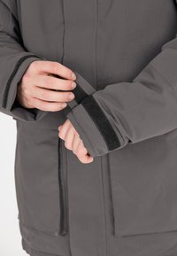 Grå vandtæt jakke med en blød tekstur, justerbare Velcro-manchetter og lige snit, med synlige sømme og sidelommer.