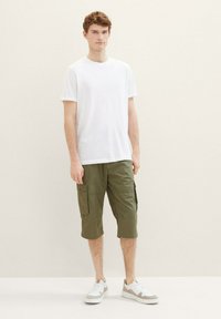 Weißes Baumwoll-T-Shirt, olivgrüne Cargo-Shorts mit Seitentaschen und graue/weiße Sneakers. Das Model steht vor einem hellen Hintergrund.