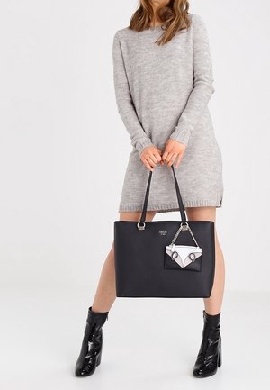 Vrouw in een grijze trui-jurk met lange mouwen, die een grote zwarte handtas vasthoudt met een klein decoratief tasje, en zwarte enkellaarsjes draagt.