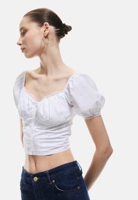 Weißes, verkürztes Oberteil mit Puffärmeln, Spitzenbesatz und Knopfverschluss; kombiniert mit dunkelblauen, hochgeschnittenen Jeans. Glatte Stofftextur.