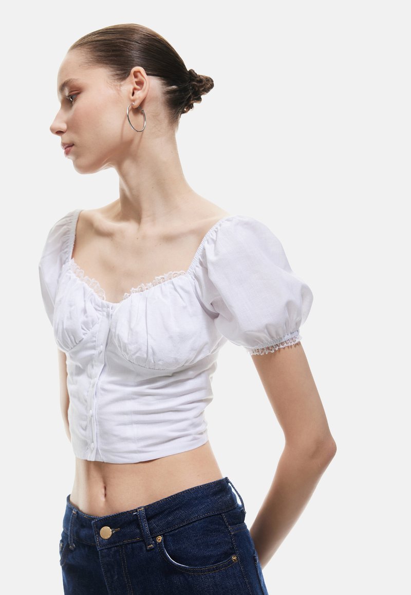 Weißes, verkürztes Oberteil mit Puffärmeln, Spitzenbesatz und Knopfverschluss; kombiniert mit dunkelblauen, hochgeschnittenen Jeans. Glatte Stofftextur.