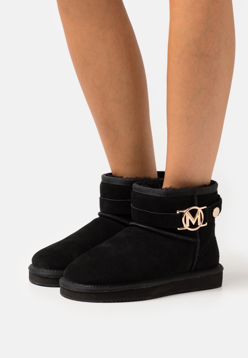 Bottines en daim noir avec un accent logo circulaire en ton or et une doublure en peluche. Tige courte avec semelle en caoutchouc plate et détails cousus.