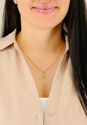 Collana d'oro con un piccolo ciondolo dettagliato che assomiglia a un orsetto. Indossata sopra una camicia beige chiaro. Design della catena semplice.