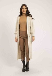 Long gilet en tricot de couleur crème avec des poches, porté par-dessus un col roulé marron et un pantalon fitted marron. Des bottines noires à la cheville complètent l'ensemble.