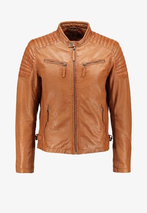 Mann trägt eine hellbraune Lederjacke, graues Hemd, schwarze Hose und schwarze Stiefel und steht mit einer Hand in der Tasche vor weißem Hintergrund.