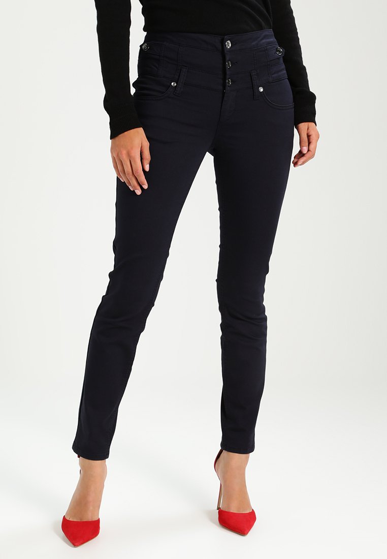 Dunkelblaue, eng anliegende Jeans mit hohem Bund, die über mehrere Knopfverschlüsse, seitliche Taschen und kontrastierende rote Pumps verfügen.