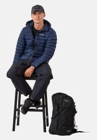 Marineblauwe pufferjack met een gequiltde textuur, zwarte cargo pants, zwarte schoenen en een zwarte rugzak die op een kruk is geplaatst. De rugzak heeft ritsen en banden.