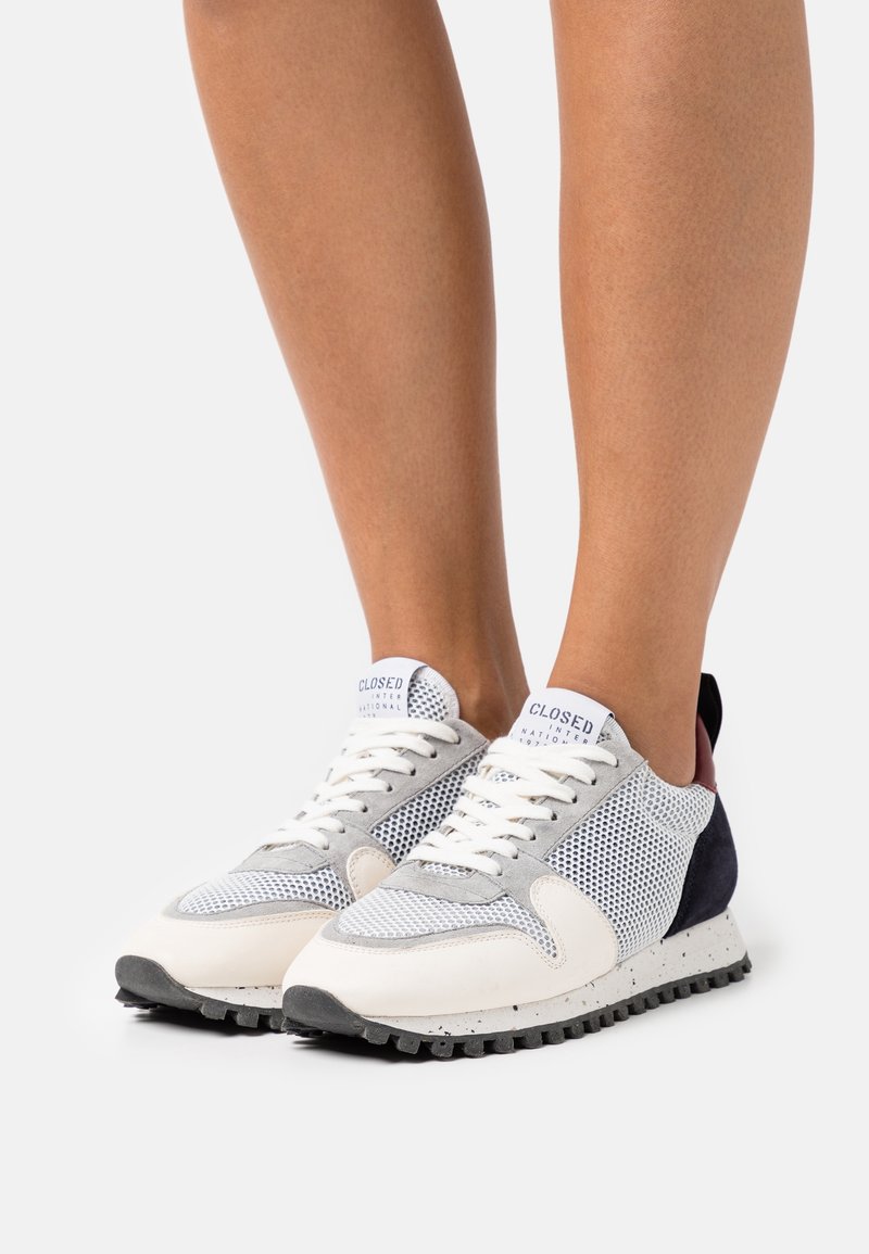 Personne portant des baskets en maille blanche et grise avec des lacets blancs et des semelles mouchetées sur un fond clair uni.