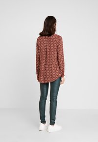 Blusa de manga longa vermelha com padrão, de corte descontraído, combinada com calças de pele sintética verdes e ténis brancos. Vista traseira mostrada.