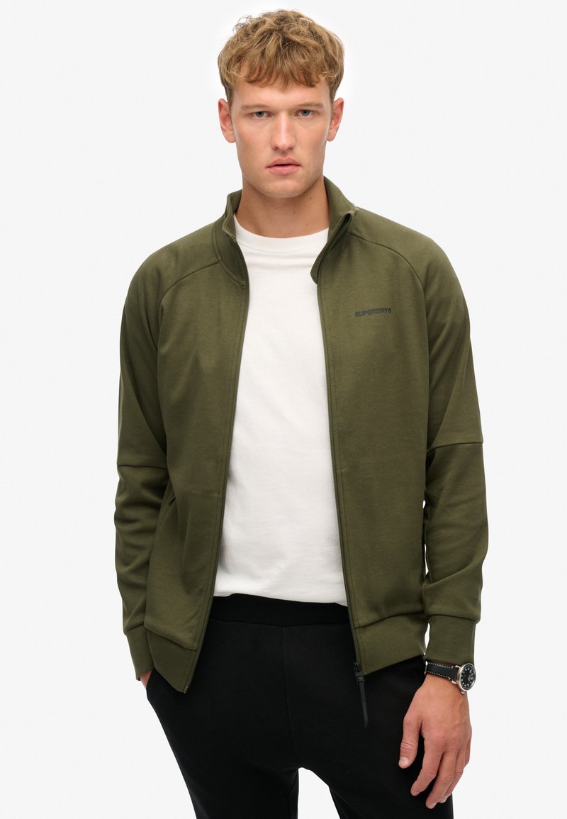 Olivengrøn zip-up jakke lavet af blødt materiale, med en slim fit, høj krave og diskret logo detalje på brystet.