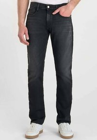 Jean en denim noir à coupe ajustée, avec cinq poches et une fermeture par bouton, porté avec des baskets blanches. Le jean présente une légère décoloration.