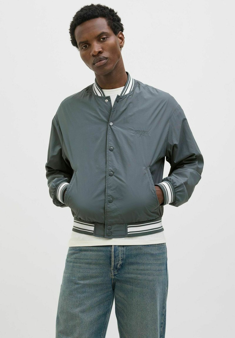 Graue Bomberjacke mit glattem, glänzendem Finish, gerippten schwarz-weißen Bündchen und Knopfverschluss. Über einem weißen Hemd getragen.