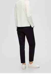 Person som bär en vit långärmad topp, svarta cropped byxor och vita sneakers, stående med händerna i fickorna mot en slät bakgrund.