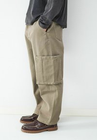 Beige cargobroek met een losse fit, grote zijzak en subtiele stikdetails. Draagt met donkerbruine, gestructureerde veterschoenen.