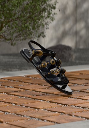 Must läikiv sandaal kuldsete naelte ja pandlatega puidust ja kivikestega mustrilisel pinnal loomulikus valguses.