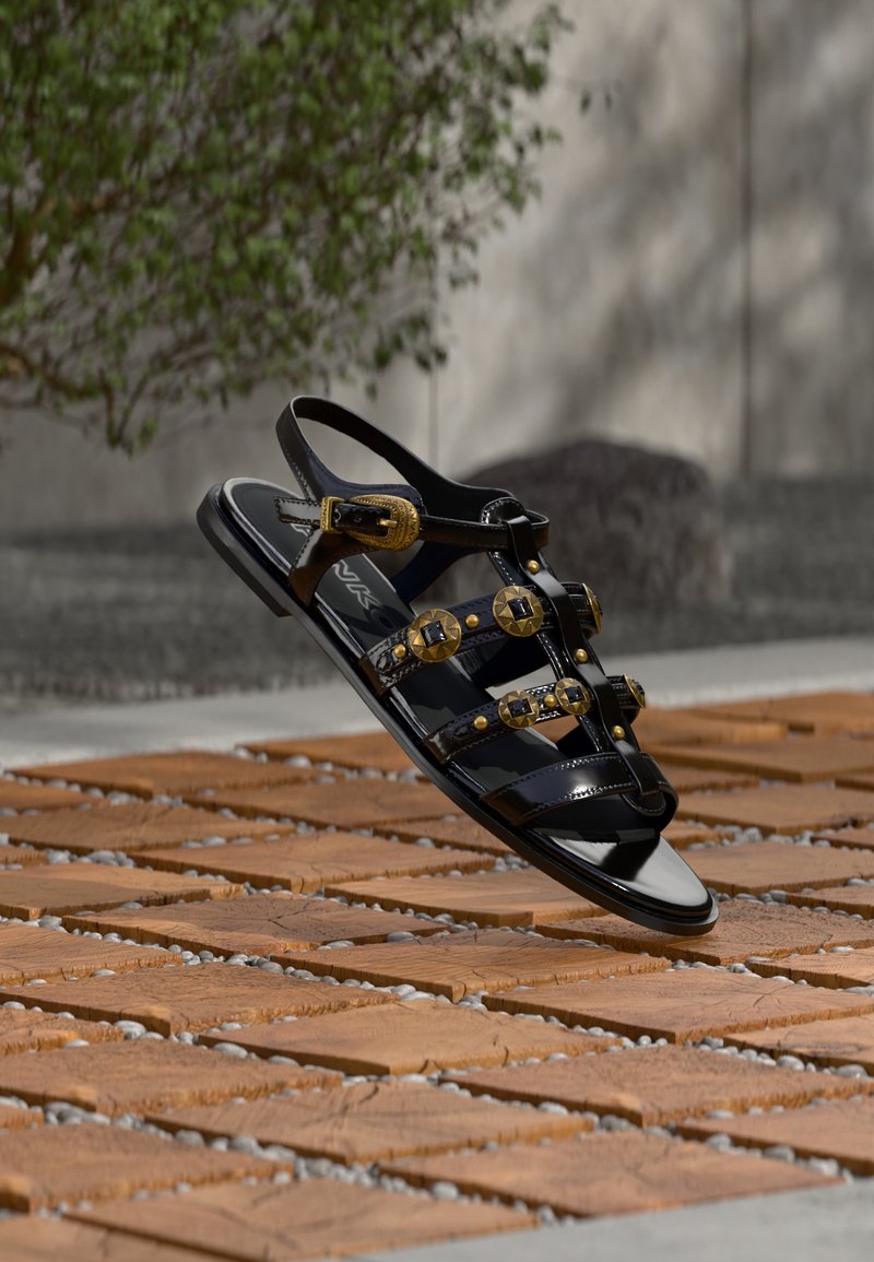 Sandale noire brillante avec des clous et boucles dorés, posée sur un sol en bois et galets sous une lumière naturelle.