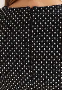 Tissu noir avec un motif géométrique en losanges blancs. Équipé d'une fermeture éclair discrète à l'arrière, offrant un design épuré et fonctionnel.