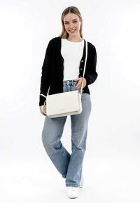 Weißes Leder-Crossbody-Bag mit strukturierter Form, silbernen Hardware-Elementen und glatter Textur. Modell trägt eine schwarze Strickjacke über einem weißen Oberteil.