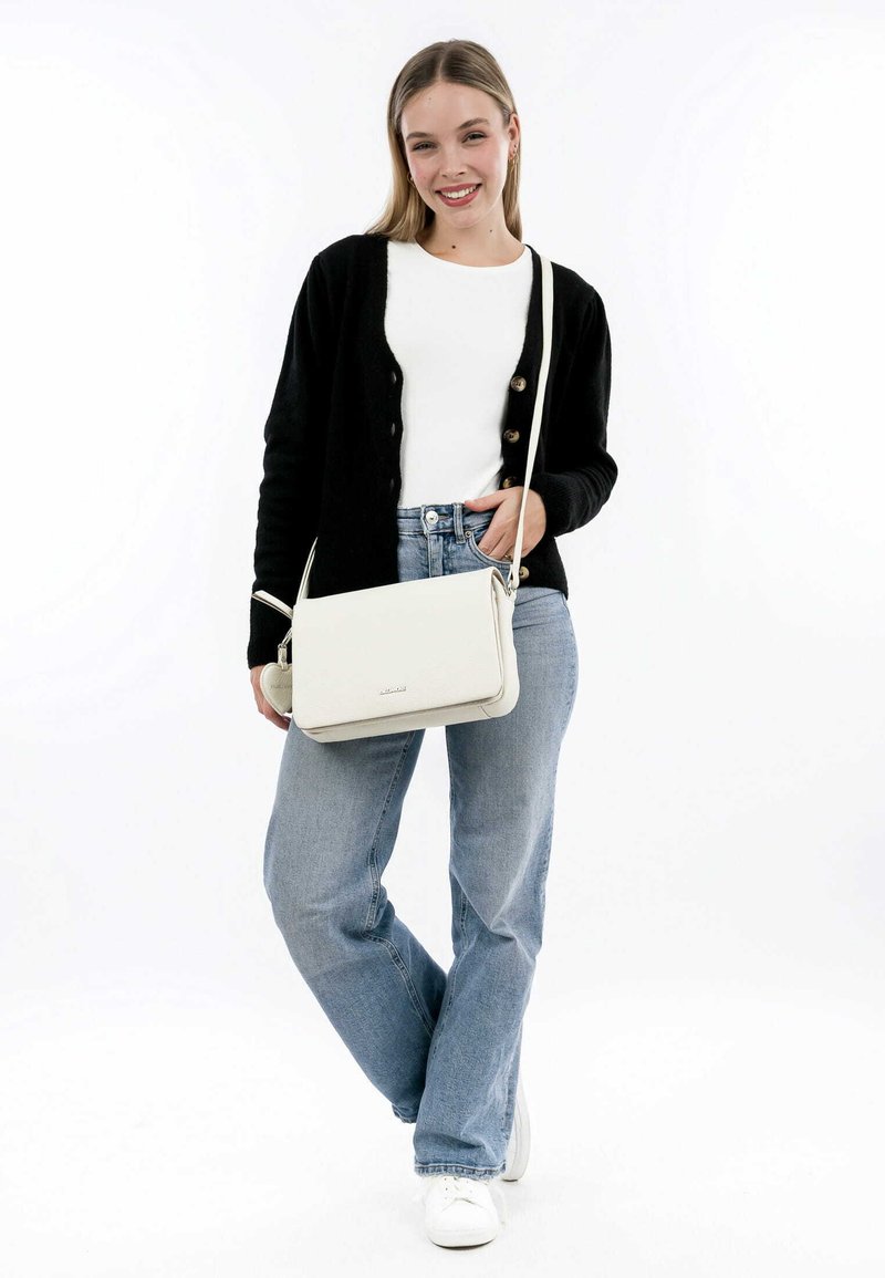 Weißes Leder-Crossbody-Bag mit strukturierter Form, silbernen Hardware-Elementen und glatter Textur. Modell trägt eine schwarze Strickjacke über einem weißen Oberteil.