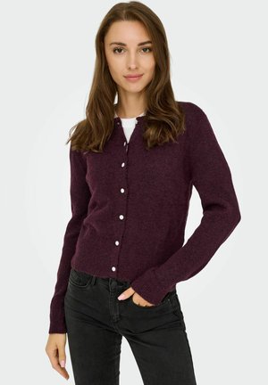 Cardigan en tricot bordeaux avec un col en V, des manches longues et des fermetures à boutons en perles. Porté sur une chemise blanche, associé à un pantalon noir.
