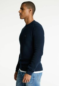 Navyblauer Pullover aus strukturierter Strickware, mit Rundhalsausschnitt, gerippten Bündchen und einem dezenten Logodetail auf der Brust.