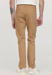 Pantalons en coton beige avec une coupe droite, deux poches arrière et une étiquette à la taille. Portés avec des baskets de couleur claire.