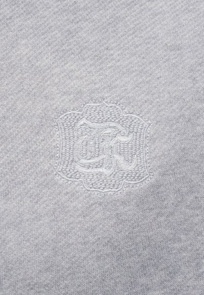 Ανοιχτό γκρι ύφασμα με ένα διακριτικό κεντητό έμβλημα που απεικονίζει στιλιζαρισμένα γράμματα μέσα σε μια διακοσμητική ασπίδα.