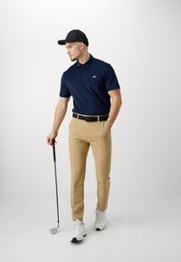 Polo shirt bleu marine, pantalon khaki, ceinture noire, chaussures de golf blanches. Le mannequin tient un club de golf et porte une casquette noire. Design minimaliste.