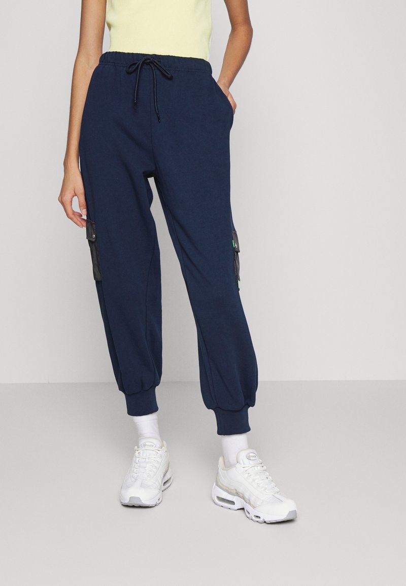 Personne debout à l'intérieur portant un pantalon de jogging bleu marine à taille à cordon, poches latérales noires, chaussettes blanches, baskets blanches et un haut jaune clair.