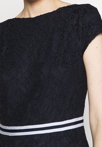 Černé krajkové šaty s krátkými rukávy a páskem v bílém a černém pruhu u pasu, zobrazené na horní části těla osoby.