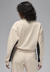 Sweatshirt de sport beige avec une bande latérale noire, coupe décontractée, poignets côtelés et col rond. Texture lisse avec des détails de couture subtils.