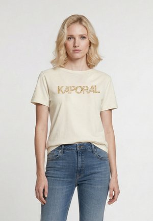 Femme blonde portant un t-shirt crème avec le texte "KAPORAL" et un jean bleu taille haute, debout devant un fond clair uni.