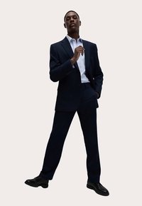 Marineblauer Anzug mit strukturiertem Design, ausgestattet mit einem Reverskragen, kombiniert mit einem hellblauen Hemd mit Knöpfen und schwarzen Schuhen.