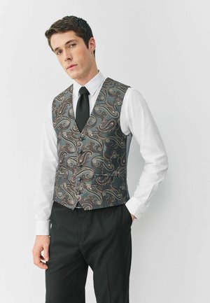 Šedý vestový kabátek s vzorem paisley a jemným leskem, odpovídající černá kravata a bílá košile, doplněné černými kalhotami.