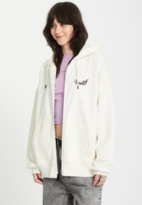 Felpa bianca con zip, dotata di una grande tasca frontale, cappuccio con coulisse e grafica nera a forma di ali sul petto. Indossata sopra un crop top lavanda.