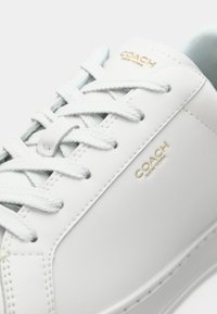 Άσπρο δερμάτινο αθλητικό παπούτσι με επίπεδα γκρι κορδόνια, χρυσή επιγραφή "COACH", στρογγυλεμένη μύτη και λεία υφή. Διαθέτει μινιμαλιστικό σχεδιασμό.