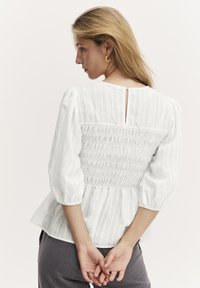 Vit blus med puffärmar, smockad rygg och nyckelhålsöppning. Texturerat randigt mönster förstärker designen. Tillsammans med grå byxor.