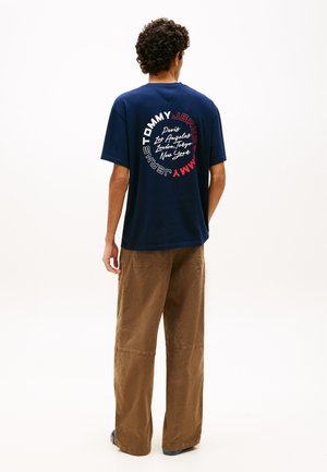 Homme portant un T-shirt bleu marine Tommy Jeans avec des noms de villes et un logo circulaire au dos, associé à un pantalon marron ample et des baskets.