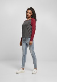 Graues Langarmshirt mit maroonfarbigen Raglanärmeln, kombiniert mit hellblauen Skinny-Jeans und weißen Sneakern vor einem einfarbigen Hintergrund.