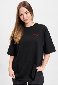Black oversized t-shirt in cotton fabric featuring a red logo on the chest. Short sleeves and a round neckline, with a relaxed fit.