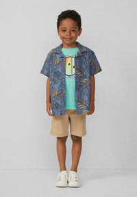 Chemise à manches courtes bleue avec des motifs de feuilles colorés, superposée à un T-shirt bleu clair avec un graphique de soleil, accompagnée de shorts beige et de baskets blanches.